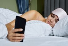Higiena cyfrowa dla zdrowia ogólnego ile ekranów to za dużo i jak ograniczyć ich wpływ na sen Kobieta w łóżku z opaską na oczach przegląda smartfon przed snem