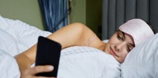 Higiena cyfrowa dla zdrowia ogólnego ile ekranów to za dużo i jak ograniczyć ich wpływ na sen Kobieta w łóżku z opaską na oczach przegląda smartfon przed snem